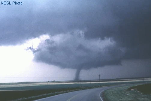 A dark, rounded lowering hangs beneath a storm base over open farmland with a narrow cone-shaped tornado beneath it. To the left of the lowering the cloud base is visibly brighter and higher, with a wedge of lighter sky cutting in toward the tornado
